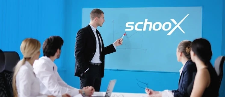 A man in a suit presents on a screen with the word Schoox, while four people watch.