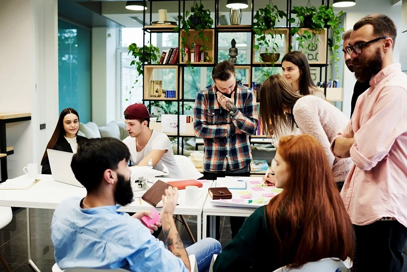 A group of six people in a modern office discussing ideas around a table with notebooks and colorful sticky notes.