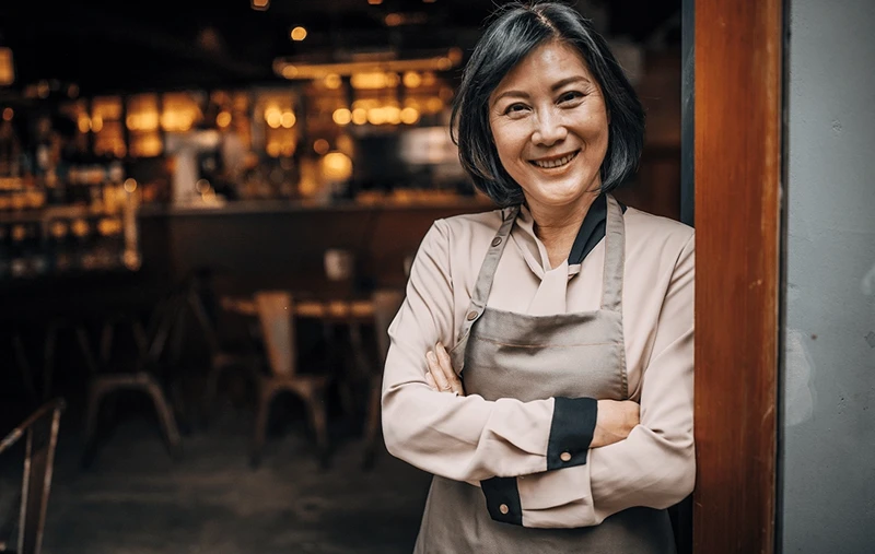 A smiling woman in a beige blouse and apron stands with arms crossed in a cafe.