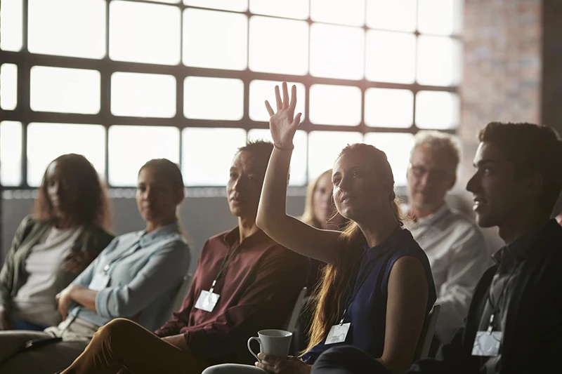 A group of people in a meeting, with a woman raising her hand to ask a question.