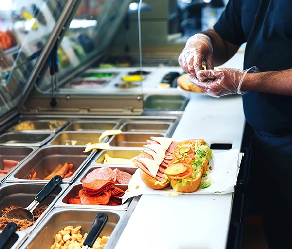 A sandwich being prepared with ham, turkey, lettuce, and tomato at a deli counter.