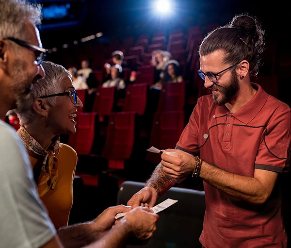 A young man gives tickets to two smiling people in a movie theater.