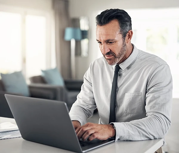 A man in a gray shirt and tie working on a laptop in a bright, modern room.