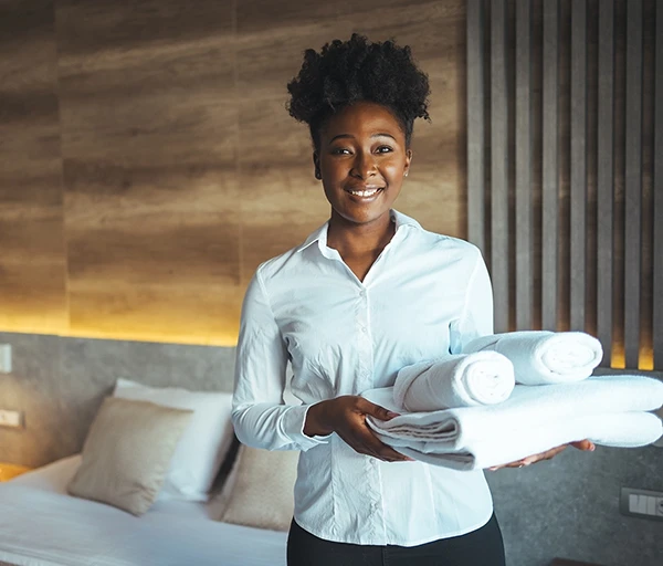 A smiling woman in a white blouse holding three rolled towels, standing in a stylish hotel room.