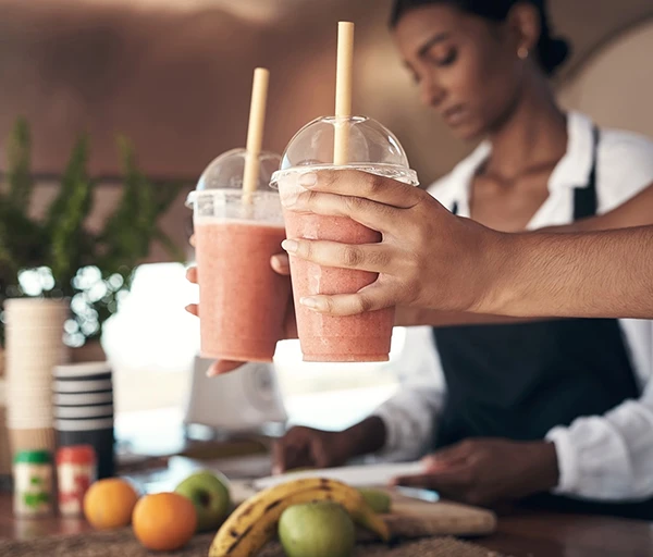 Two hands holding cups of pink smoothies with yellow straws, while a woman prepares food behind.
