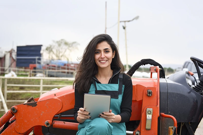 A smiling woman in overalls sits on a tractor holding a tablet, surrounded by farmland.