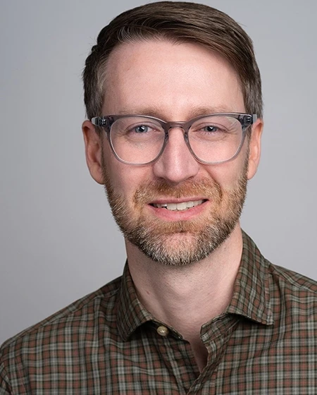 A smiling man with glasses and a beard, wearing a checkered shirt.