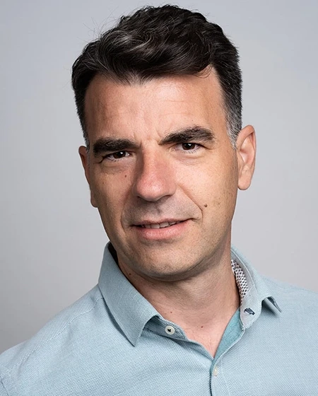 A man with dark hair wearing a light blue shirt, smiling at the camera.