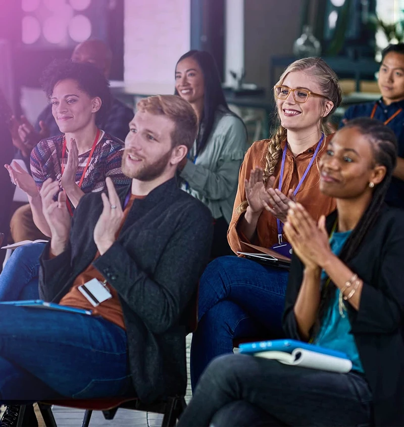 A diverse group of people seated, clapping in a bright meeting space.
