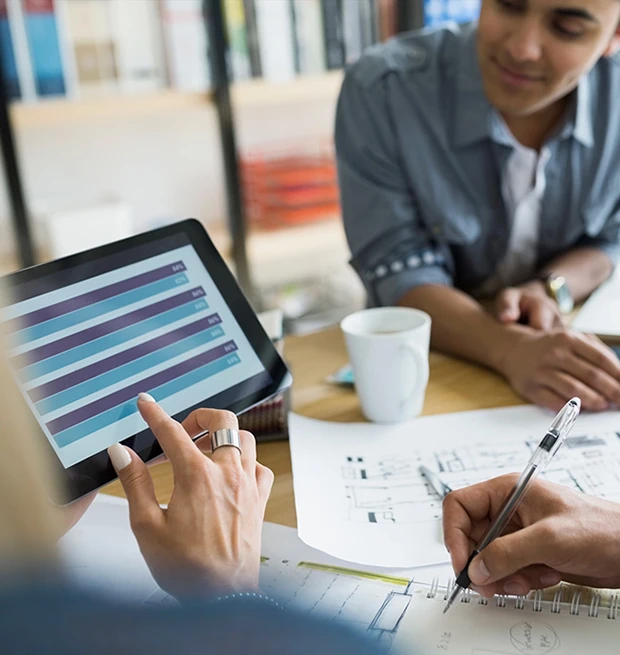 A person holds a tablet showing a chart while discussing project plans with another person, with coffee and sketches on the table.