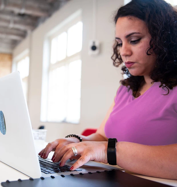 A woman in a purple shirt typing on a laptop at a bright workspace.