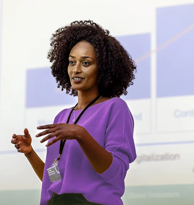 A woman with curly hair in a purple sweater gives a presentation, gesturing with her hands.