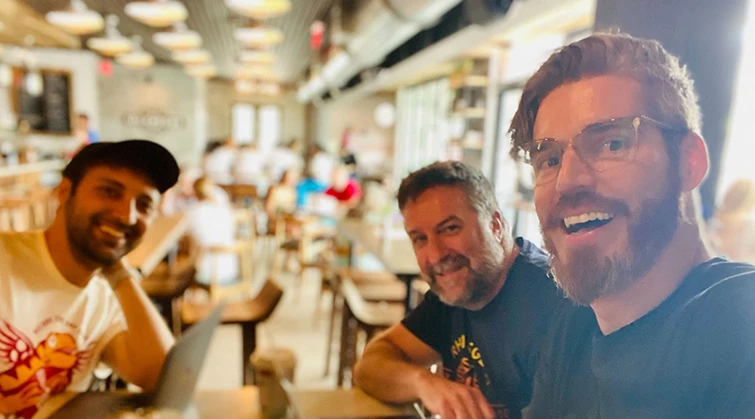Three smiling men sitting at a table in a busy cafe, with chairs and patrons in background.