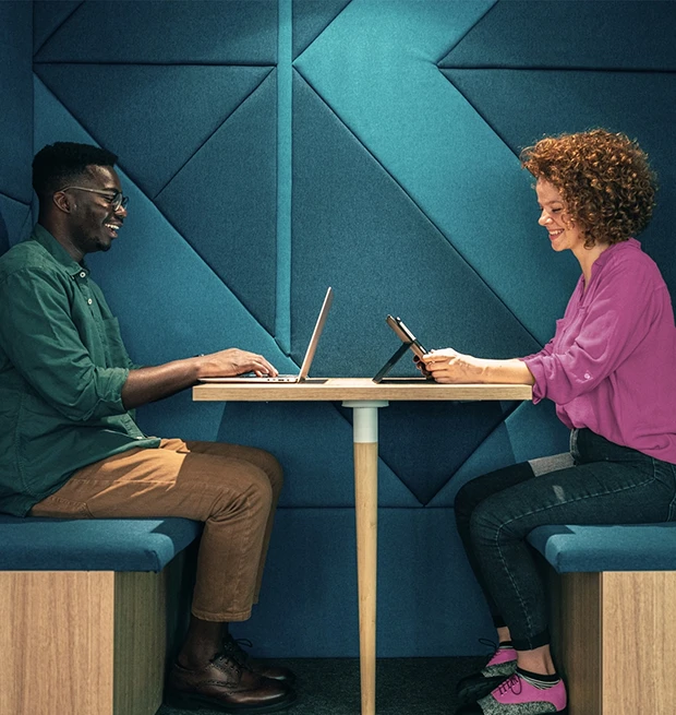 Two people seated at a table, one using a laptop and the other a tablet, smiling at each other.