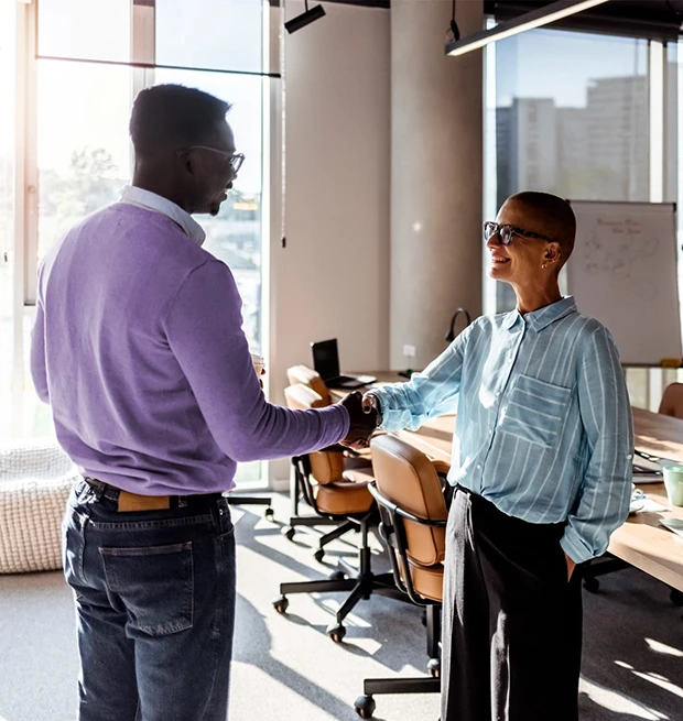 A man shakes hands with a woman in a modern office, sunlight streaming through windows.