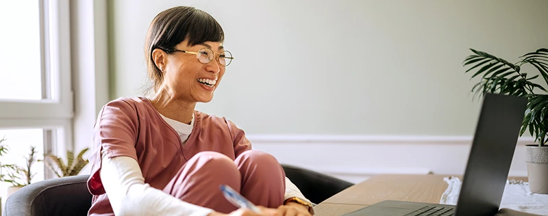 A smiling woman in scrubs sits at a table using a laptop, with plants in the background.