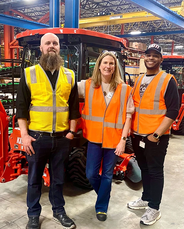 Three people in safety vests pose in a warehouse with red tractors in the background.