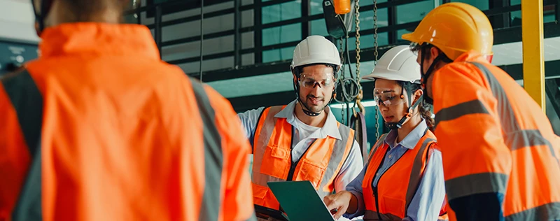Three construction workers in safety gear discuss plans in a warehouse setting.