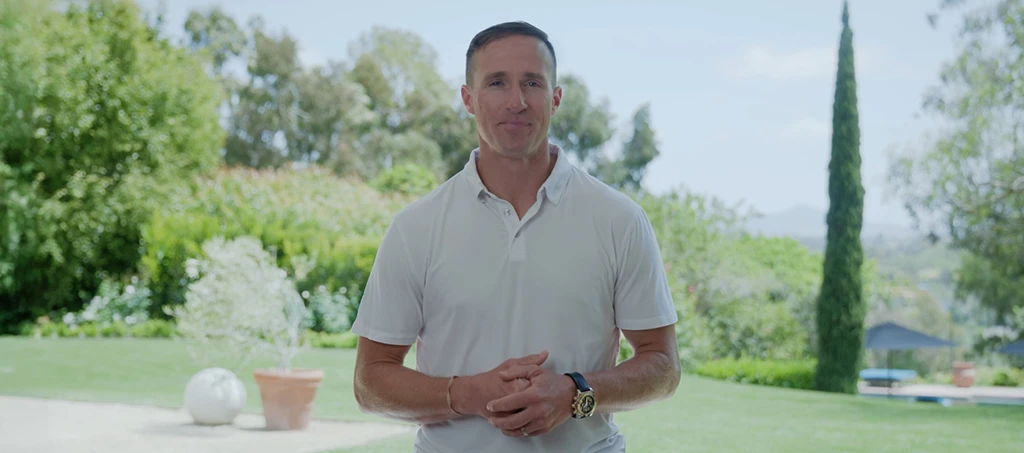 Drew Brees stands outdoors in a white polo shirt, smiling in a garden with trees and a pool in the background.