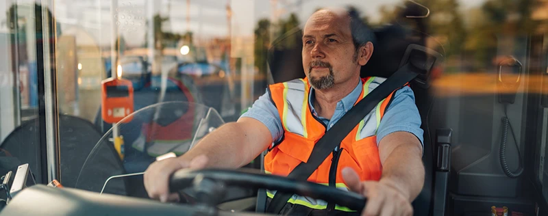 A bus driver in an orange vest focuses on the road while driving a bus.