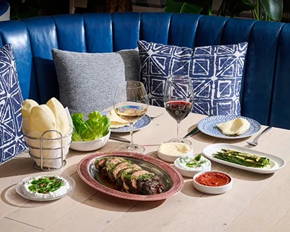 A table set with grilled meat platter, sauces, lettuce, pita bread, and glasses of wine.