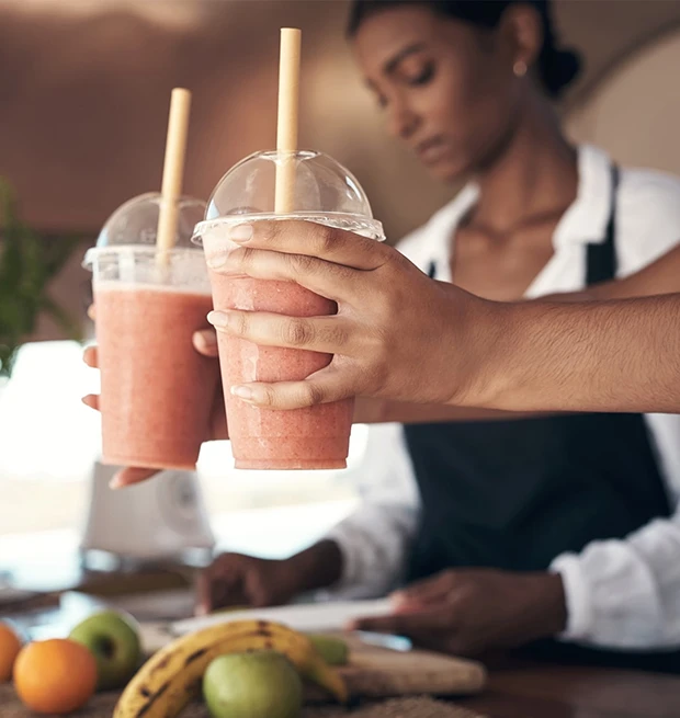 Two hands holding fruit smoothies with straws in a café, a woman is preparing in the background.