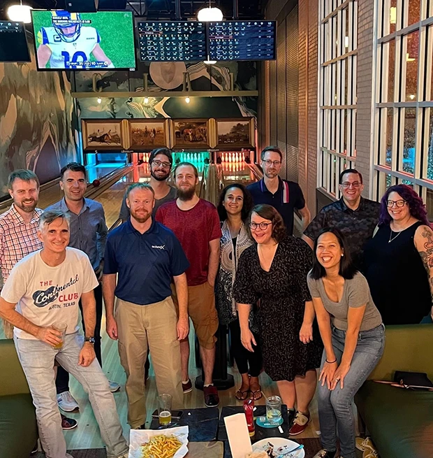 A group of 15 people smiling together at a bowling alley, with pins and a scoreboard behind them.