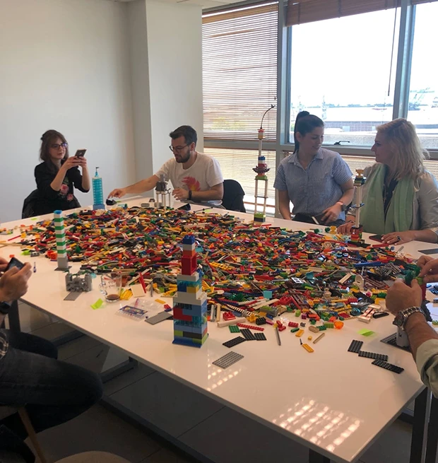 A group of people building with colorful LEGO pieces on a white table in a bright room.