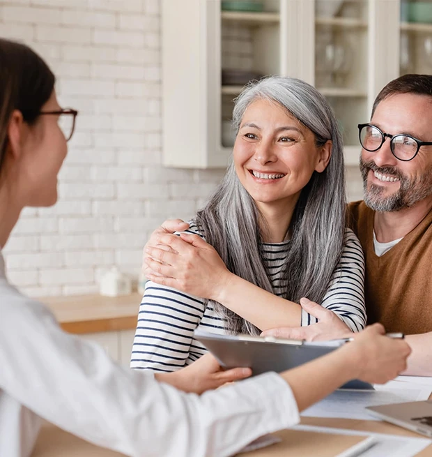 A happy couple with gray hair embraces while talking to a woman holding a tablet.