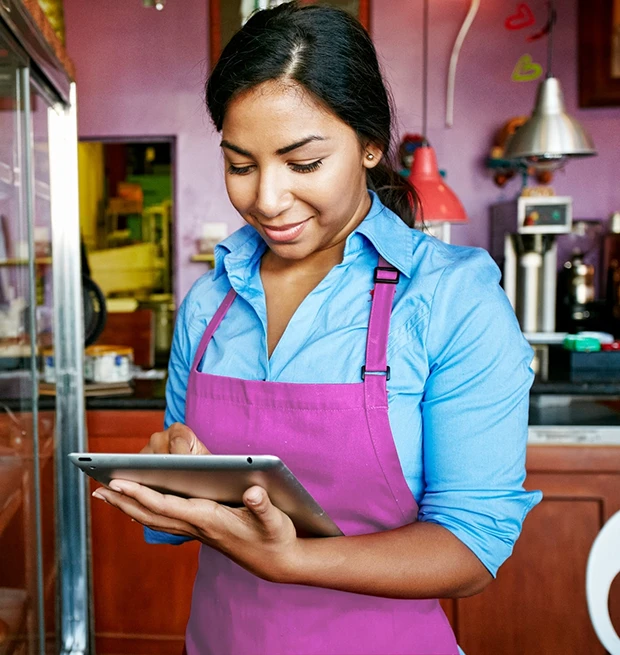 A woman in a blue shirt and pink apron using a tablet in a cozy cafe.