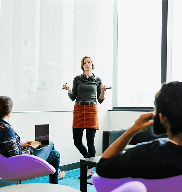 A woman stands in front of a whiteboard, explaining something to seated colleagues in a modern office.