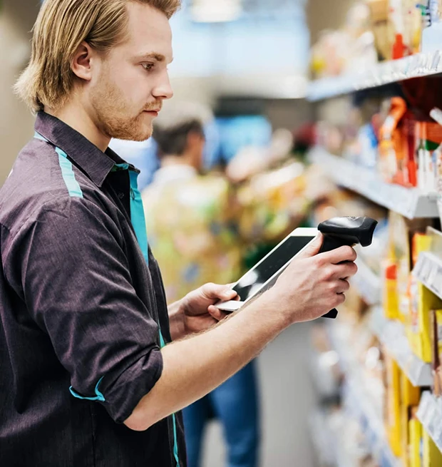 A young man scans items on a store shelf using a tablet and barcode scanner.