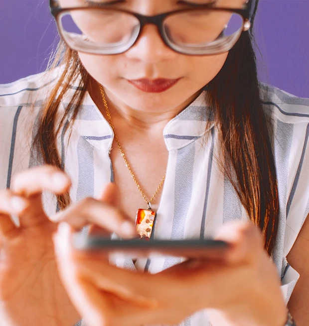 A woman holding a smartphone while wearing glasses and a necklace, focused on her screen.