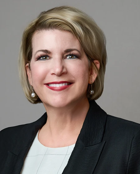 A smiling woman with short blonde hair, wearing a black blazer and pearl earrings, against a gray background.