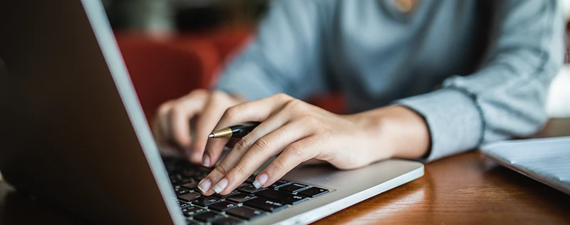 A person typing on a laptop with a pen in hand, sitting at a wooden desk.