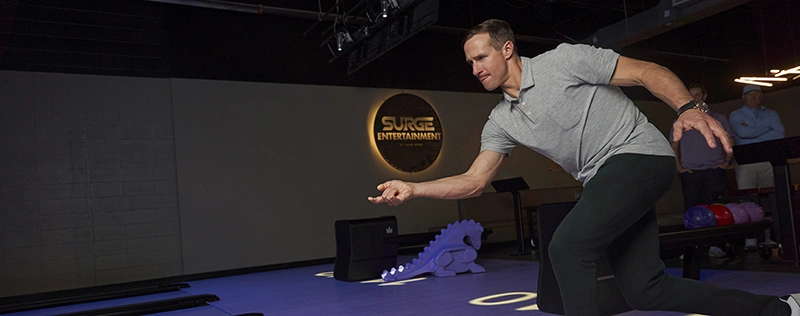 A man in a gray shirt leans forward to throw a ball at a bowling lane in a dark entertainment venue.