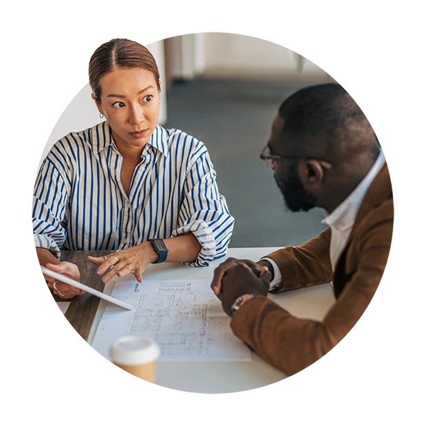 A woman and a man discuss plans at a table, with blueprints in front of them.