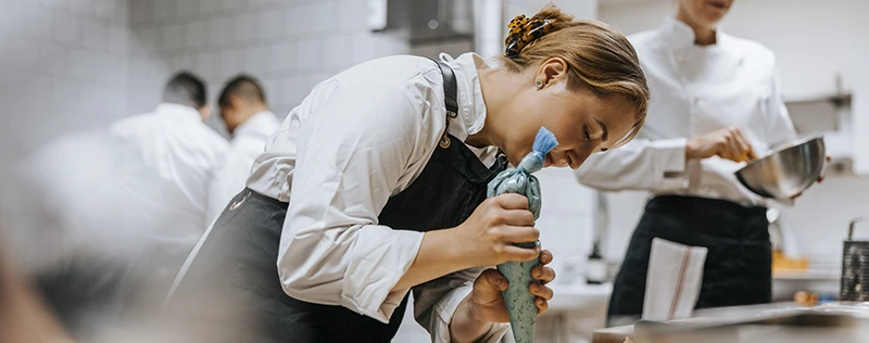 A chef in an apron carefully squeezes a blue piping bag in a busy kitchen.