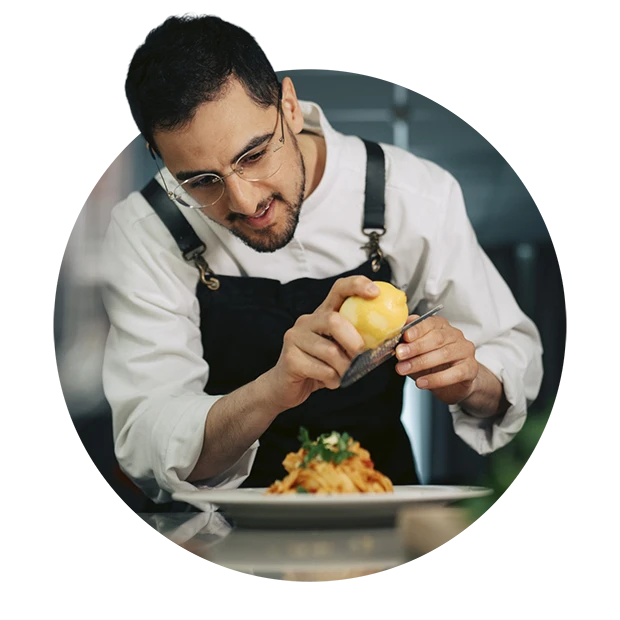 A chef grating lemon zest over a plate of pasta garnished with herbs.