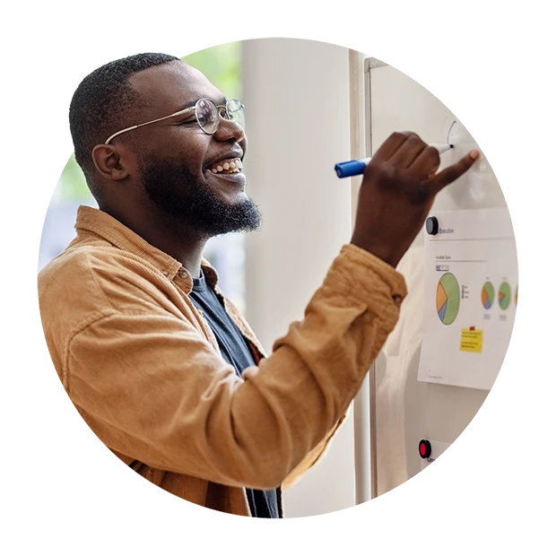 A smiling man with glasses is writing on a whiteboard, surrounded by colorful graphs.