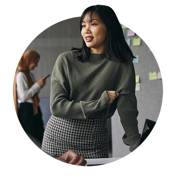 A woman in a green sweater and checked skirt smiles while talking, with sticky notes on a wall behind her.