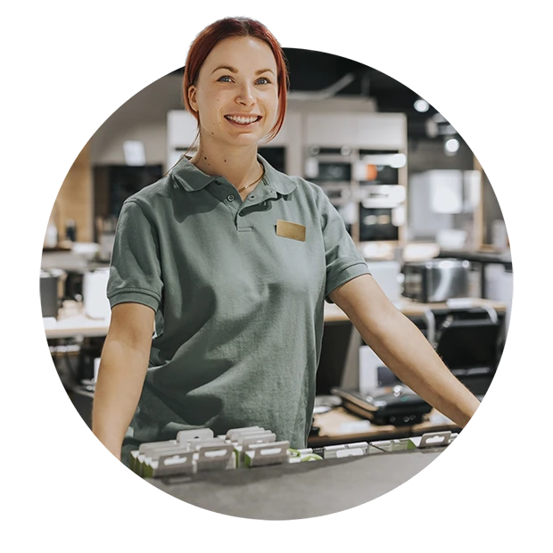 A smiling woman with red hair in a green polo shirt stands at a store counter.