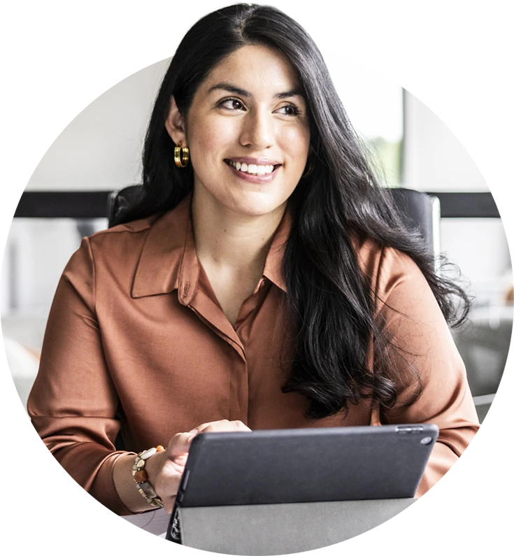 A woman with long dark hair, wearing a brown shirt, smiles while using a tablet at a table.