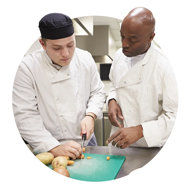 A chef teaching a student how to chop vegetables on a green cutting board.