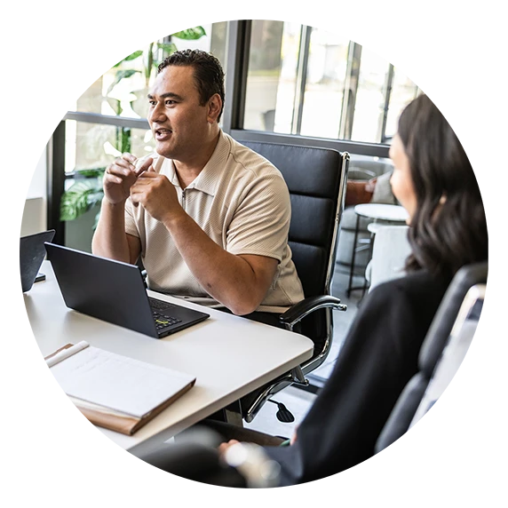 A man in a beige shirt gestures while talking at a meeting table with a laptop.