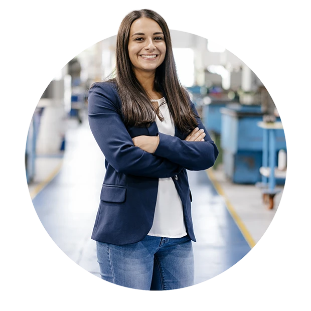 A woman in a black blazer and white top stands confidently with arms crossed in a factory.