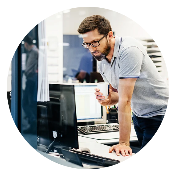 A man in a gray polo shirt, looking at a computer screen while holding a cup.