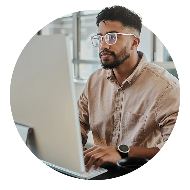 A man with glasses working on a computer, wearing a beige shirt, focused and seated at a desk.