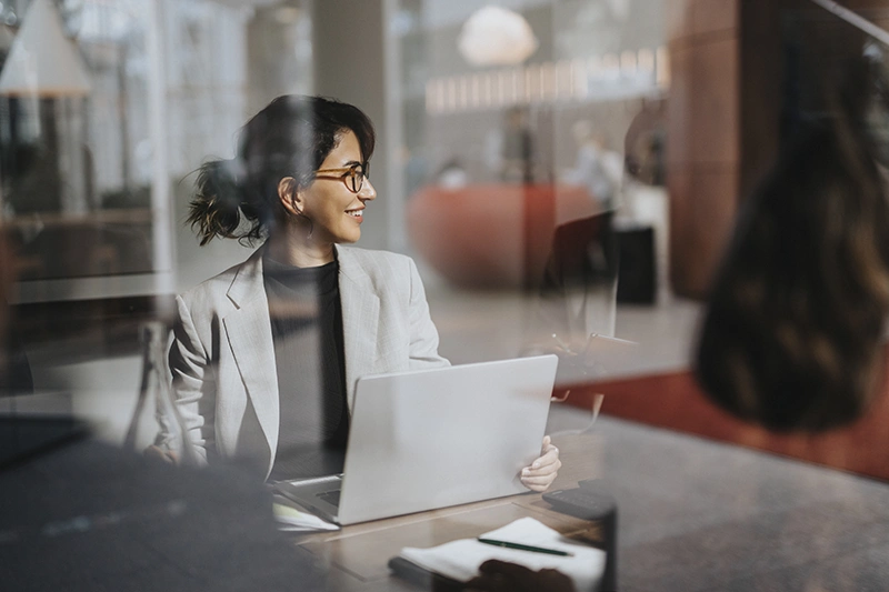 A smiling woman with glasses sitting at a table with a laptop, working in a bright office.