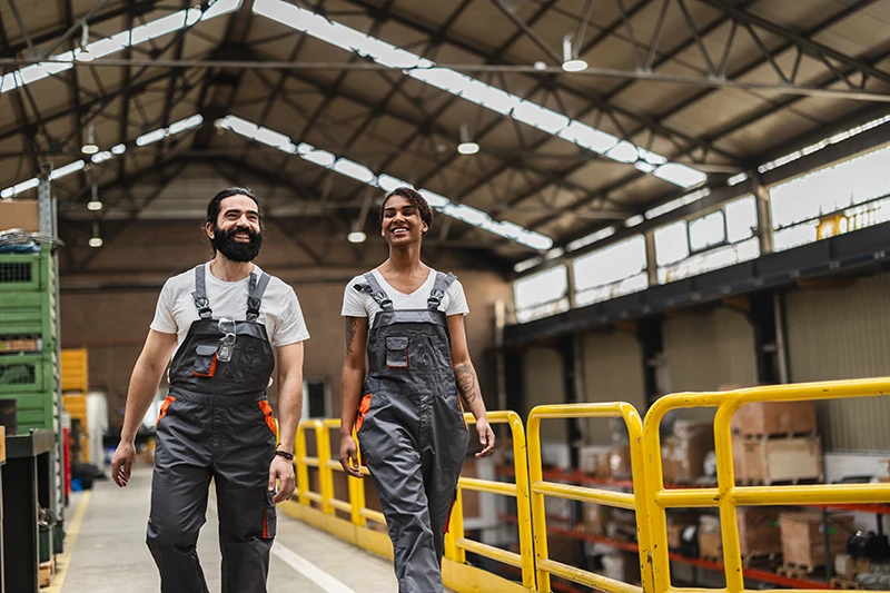 A man and woman in gray overalls walk together in a bright warehouse.